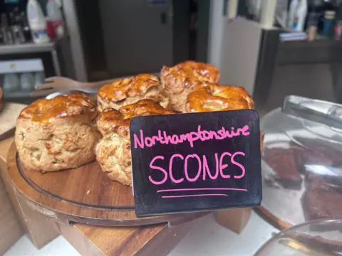 Four freshly baked scones on a plate. There is a hand-written sign on a slate saying "Northamptonshire Scones"