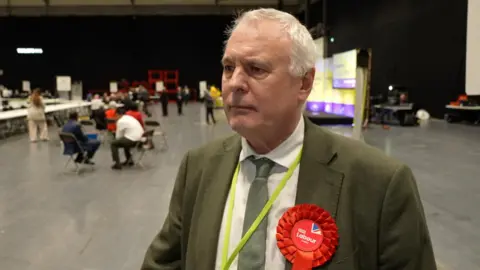 George Duggins at the Coventry City Council election count. He wears a green jacket and tie and a Labour rosette.