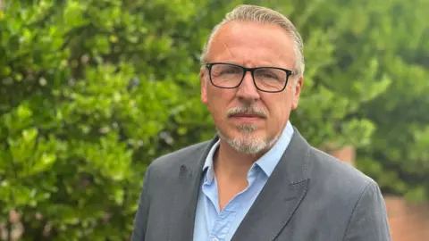 A man is stood looking towards the camera. He is wearing glasses, has grey hair and facial hair. He is wearing a blue shirt with a suit jacket over the top. He is stood in front of a green bush and a brown wall