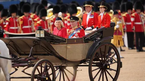 Getty Images The King and Queen riding in a carriage. He is wearing the uniform of the Grenadier Guards as well as a number of medals. Queen Camilla is wearing a red, silk crepe, grenadier guards uniform dress and a black beret. Rows of troops stand behind them.