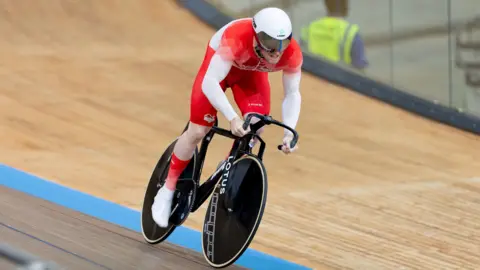 PA Media A man wearing a red cycling suit and white helmet is seen riding on an indoor cycling track. His face is obscured by a visor, and there is a glass screen to his side at the edge of the track.