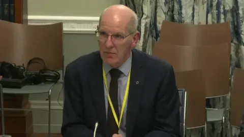 A bald man in a navy suit sits at a table. He is wearing a grey shirt with dark tie and yellow lanyard. There is a silver circle pin (fáinne) on his right lapel. 