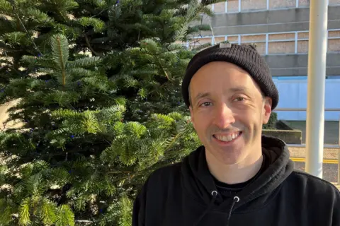 John Devine/BBC Rob, a man, wearing a black woollen hat and a black hoodie, smiling for camera. A green fur tree is behind him, and a concrete 1960s block in the background.