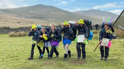 A group of six children is walking along a grass area with hills in the background. They are laughing and holding on to their yellow hats in the wind.