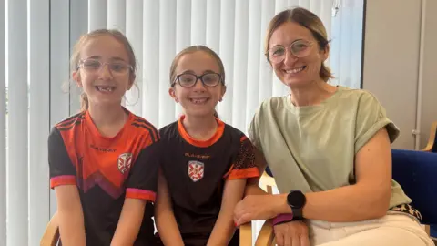 Natalie Geoghegan has dark blonde hair pulled back into a ponytail. She is wearing a light green short-sleeved top and white trousers and is sitting with two girls. She and the girls are all wearing glasses. The two girls are wearing sport tops which are black and red with a white and red logo in the middle.