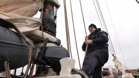 Keith Burtt pulling a rope on the ship. He is wearing all black and a black jacket. 