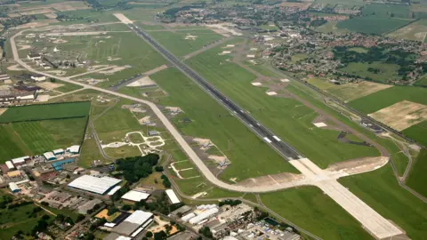 Mike Page Aerial Photogrpahy Lapangan terbang RAF Mildenhall terlihat dari udara, menampilkan landasan pacu yang dikelilingi rumput dan jalan perimeter. Kawasan industri ada di bagian bawah gambar, dengan desa Beck Row di kanan atas.