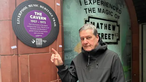 BBC Peter Hooton is dressed in a black jacket and is standing while pointing at a heritage trail plaque which reads "The Cavern".