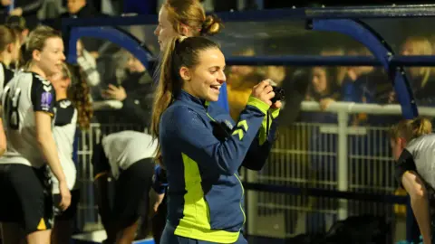 Alex - a young woman with long hair in a ponytail, and wearing navy blue and bright yellow sports kit - is holding a camera towards her team on the football pitch.