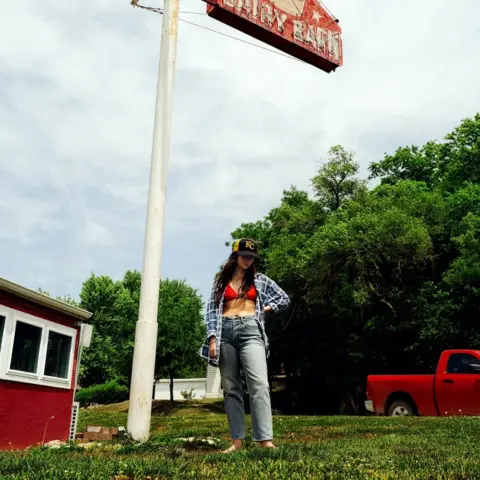 Anti- Artwork for Waxahatchee's album, Tigers Blood. A woman in jeans and an open  chequered shirt, which shows red bikini style top. She is standing on grass. In the background are trees, a red pick up and a small red building