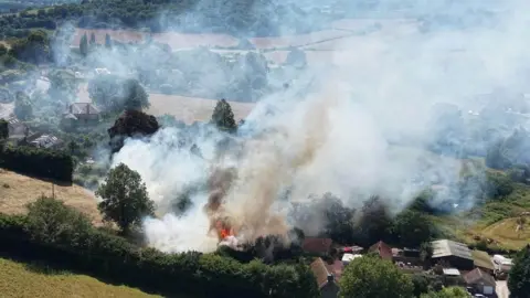 Fire and smoke can be seen rising from a rural plot of land with houses surrounded by fields and trees