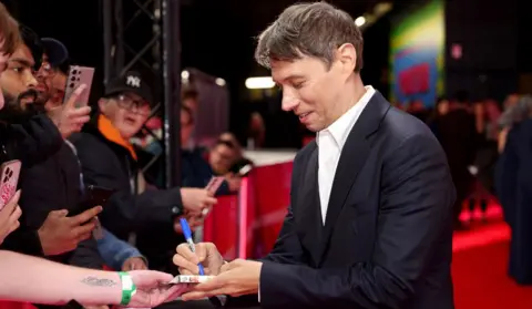 Getty Images Sean Baker attends the "Anora" Headline Gala during the 68th BFI London Film Festival at The Royal Festival Hall on October 11, 2024 in London, England. He is signing an autograph for a fan while on the red carpet.