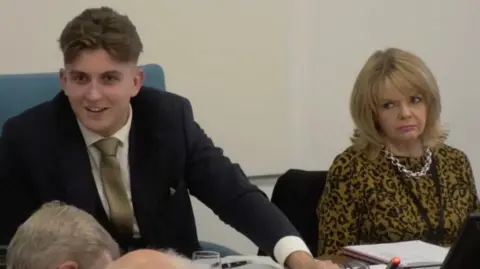 Warwickshire County Council George Finch is wearing a navy suit jacket with a gold tie and pocket square. He appears to be looking off camera at somebody. To his right Monica Fogarty, who has shoulder length blonde hair and is wearing a leopard print top and a chunky chain necklace, looks towards Finch with a frown.