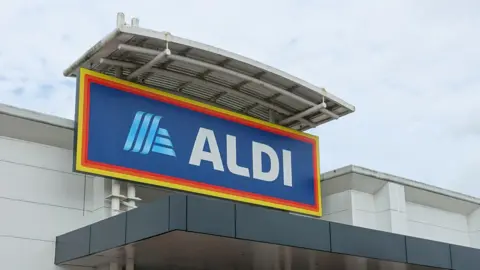 Close up of Aldi sign, which is blue with white lettering. The sign is sitting at the top of a white building with grey clouds in the background.