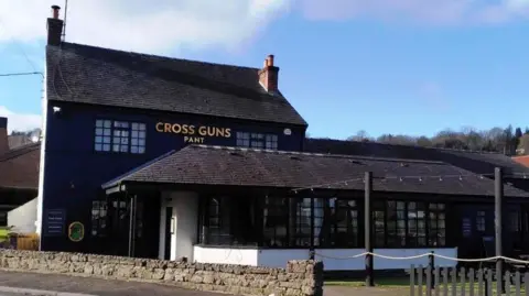 Friends of Cross Guns Pant A large dark building with a dark tiled roof and an extension at the front with the words Cross Guns Pant in gold lettering on the wall