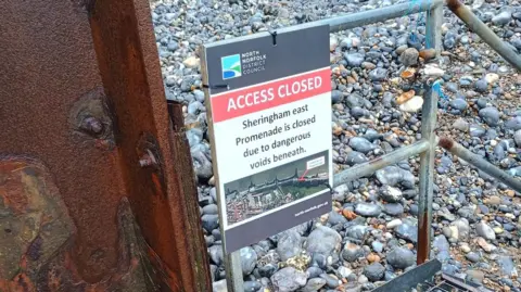 North Norfolk District Council A seafront, with an access closed sign, with metal railings, and a rusted metal structure to the left and grey pebbles on the beach. 