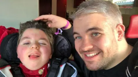 Paul Woodward, who has short grey hair and dark stubble, sits beside his son who is in a wheelchair. Both are smiling