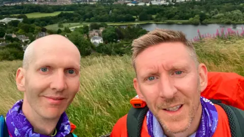 Two men smile for a picture with a green landscape and lake stretching out behind them. The man on the left is shaven, wearing a purple snood. The man on the right also wears a purple snood or scarf, wearing orange clothing.