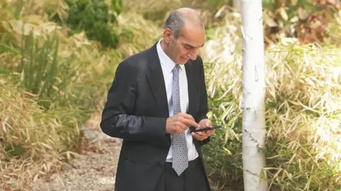 Eddie Mitchell A balding man in a dark suit, white shirt and patterned tie checks his mobile phone. 