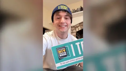 A man wearing an I Test beanie hat, smiling and holding a HIV awareness week sign.