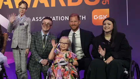 PA Media A man wearing a dark suit stands with four award winners on stage. A little girl in front of his wears a floral dress and has blonde hair and glasses. 