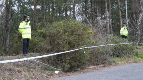 Police tape surrounding a wooded area next to a road with two officers standing on scene guard.