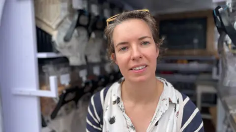 Alice Lightowlers looking at the camera. She is wearing a white shirt and white and navy striped cardigan. Behind her there are jars of dried food with dispenser nozzles. 
