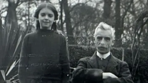 A black and white image of Elsa and William Hall. She is long dark hair pulled back from her face and is wearing a dark jumper over a dark skirt. He is sitting to her left and has white hair and a white moustache. He has a high white collar under a dark jacket and waistcoat and has his arms folded. Behind them is a garden.