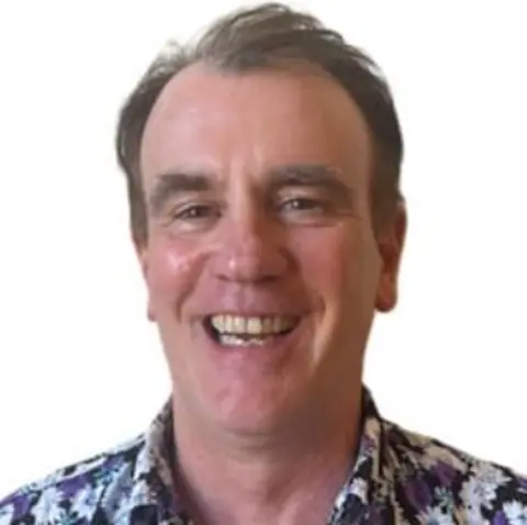 Marc Wilkinson A man with short brown hair smiles directly at the camera, wearing a blue patterned shirt.