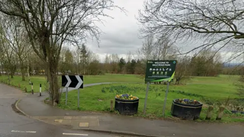 A sign which reads Dale Road Park in front of a green space, with trees in the distance and grey sky above