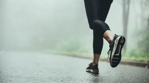 Female runner showing just her legs wearing black leggings and black and white trainers. Running on a road near grass in the fog.