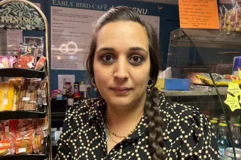 A woman with dark hair in a plait wearing a black and yellow shirt, standing behind the counter of a cafe.