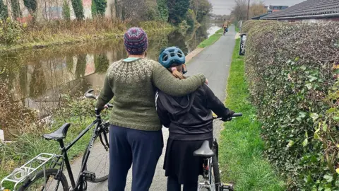 BBC A mother stands with her back to the camera with her arm around her daughter's shoulders. They are standing next to the Union Canal and both are holding their bikes. The mum is on the left and is wearing a green knitted jumper and a colourful wooly hat and the daughter is wearing a cycling helmet and dark skirt and top.