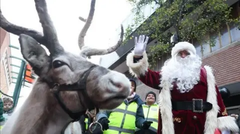 A reindeer is standing next to a waving Santa Claus during Middlesbrough Christmas parade. They are surrounded by security workers wearing high visibility vests. Several children are watching the event. 