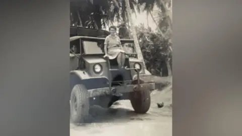 Gillian Edwards An old black and white photo showing Jane as a young woman in nurses' uniform sitting on the front of a jeep-style ambulance, smiling to camera. 