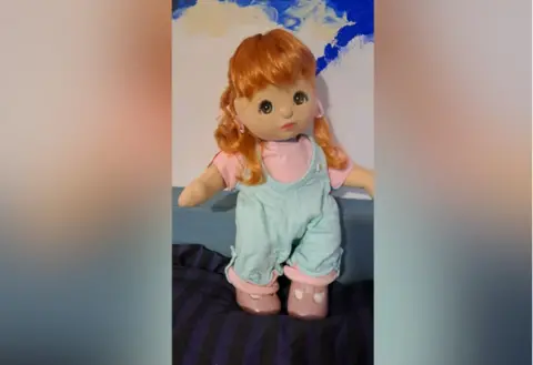 Sobell House The vintage red-haired My Girl Doll that was donated to Sobell House hospice and was sold for £310 to a buyer in Italy. It is propped up on what looks like a soft service against a wall. The doll is dressed in light blue jumpsuit, a pink T-shirt and pink shoes.