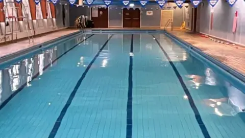 Interior of Ulverston Pool. It is a leisure pool with lanes marked with darker blue tiles at the bottom. There are flags hanging over the pool.