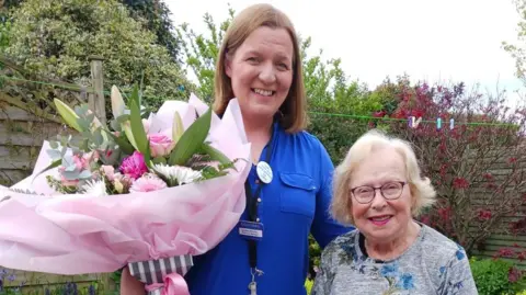 Gordons Lyndsay Sinclair has long brown hair and is wearing a blue uniform and name badge. She is holding a large bouquet of flowers in her right hand and has her other arm around Sadie. Sadie is wearing a multi-coloured top and black-rimmed glasses. 
