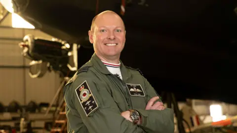 Squadron Leader Paul Wise stood wearing military uniform with his arms crossed in a warehouse in front of a military aeroplane, which is blurred in the background. He is bald and is smiling at the camera. 