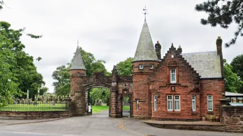 The gates of a cemetery with a red sandstone building adjoining an archway with turrets. 