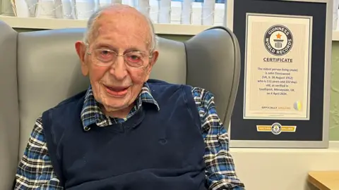 PA Media John Tinniswood, wearing glasses, a blue tank top and a blue, black and white checked shirt, sits in a grey chair and smiles at the camera. On a shelf to his left sits a framed certificate proving the Guinness World Record age achievement. 