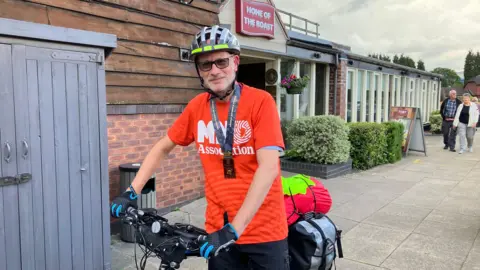 A man on a bicycle, wearing a helmet, gloves, a lanyard and a red T-shirt reading MND Association.