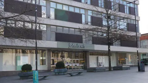 An old 1960s building has large glass windows, brown tiled panels and a white tiled facade. There is a sign above the entrance doors saying John Lewis
