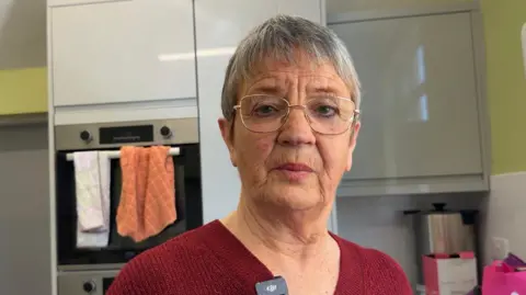 Jasmine Lowe/BBC News A lady with grey short hair. She has rosy red lip stick on which matches her red v neck jumper. she is wearing glasses with a metal frame looking at the camera. She's stood in a kitchen. 