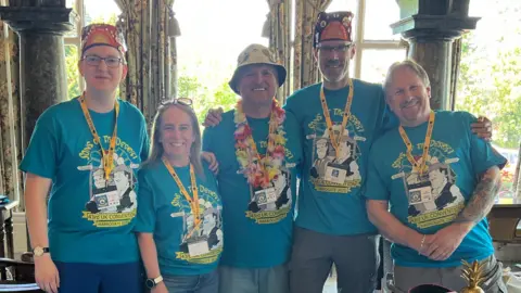 Five people in blue T-shirts stand behind a table covered in a white table cloth. Two of them are wearing fez hats and one is wearing a bucket hat.