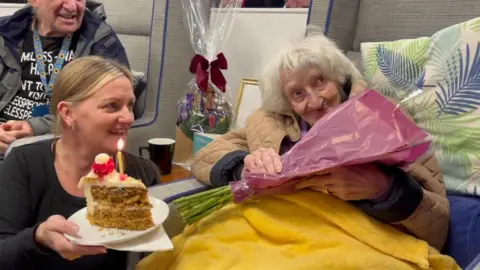 Maisie Bartlett sitting in an armchair, covered in a yellow blanket and holding a bunch of flowers. She is surrounded by well-wishers.