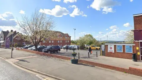 The image shows a car park next sandwiched between buildings. Several trees are growing in the car park, beginning to show leaves. A purple sign with a silver moon and the words "Premier Inn" can be seen in the left corner.
