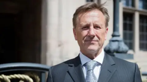 Staffordshire County Council A man with brown hair, a grey suit and pale grey tie with grey hair standing in front of a grey building