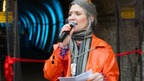 Galileo Photography A woman with greying hair wearing a bright orange coat is speaking on a microphone during a ceremony.
