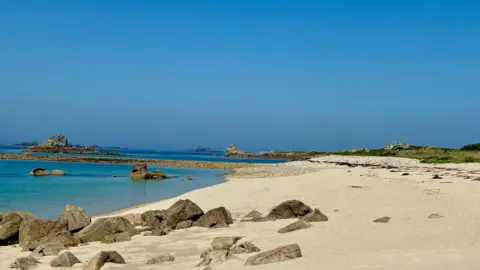 Bryher/BBC Weather Watchers Bright blue skies over the sandy beach on the Isles of Scilly. Rocks are scattered on the sand.